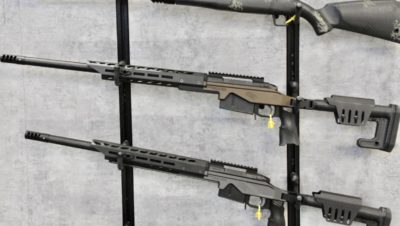 A pair of rifles rest on a wall rack. They are bronze colored with carbon fiber accents.